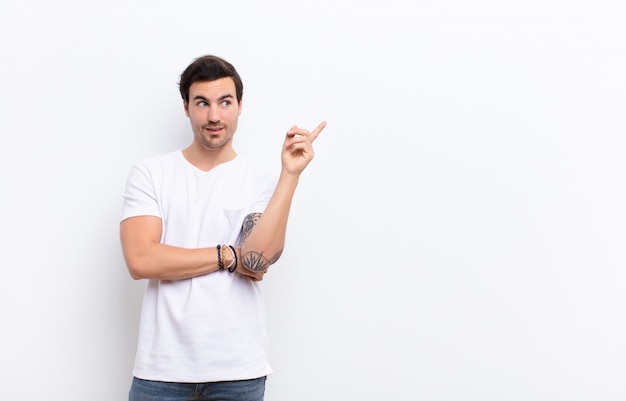 Jeune bel homme souriant joyeusement et regardant de côté, se demandant, pensant ou ayant une idée sur le mur blanc