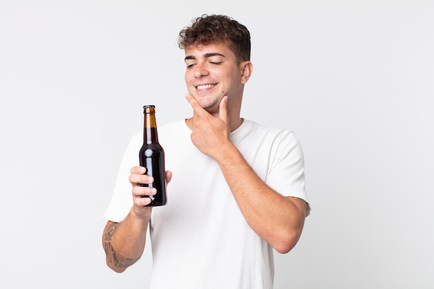 Jeune bel homme souriant joyeusement et rêvant ou doutant et tenant une bouteille de bière