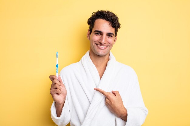 Jeune bel homme souriant joyeusement, se sentant heureux et pointant vers le côté. concept de peignoir et de brosse à dents