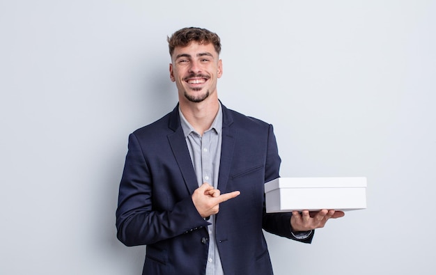 Jeune bel homme souriant joyeusement, se sentant heureux et pointant vers le côté. paquet de boîte blanche