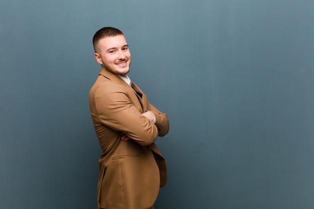 Jeune bel homme souriant joyeusement, se sentant heureux, satisfait et détendu, les bras croisés et regardant sur le côté contre un mur plat