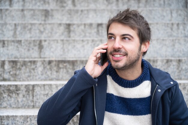 Jeune bel homme, souriant, parlant au téléphone, assis sur les escaliers.