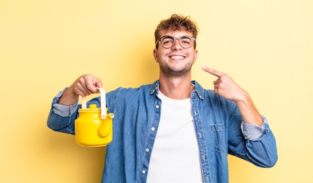 Jeune bel homme souriant pointant avec confiance vers son propre large sourire. notion de théière