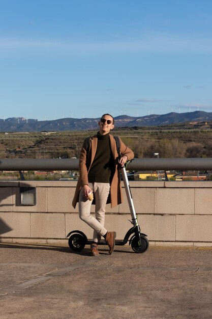 Jeune bel homme souriant profitant de la journée ensoleillée en hiver avec un café chaud et un scooter électrique pour se déplacer