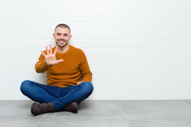 Jeune bel homme souriant et regardant amical, montrant le numéro cinq ou cinquième avec la main vers l'avant, compte à rebours assis sur le sol