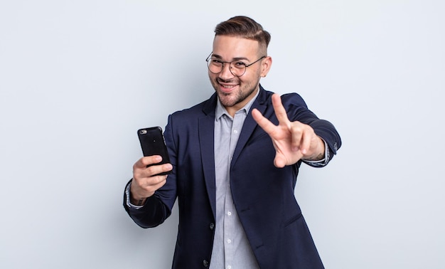 Jeune bel homme souriant et regardant heureux gesticulant victoire ou paix entreprise et concept de smartphone