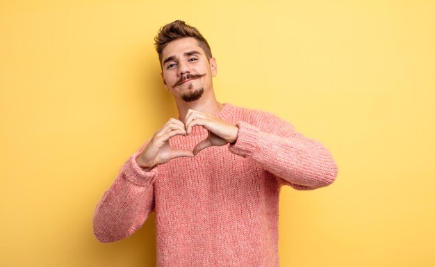 Jeune bel homme souriant et se sentant heureux, mignon, romantique et amoureux, en forme de coeur avec les deux mains. concept de moustache étrange