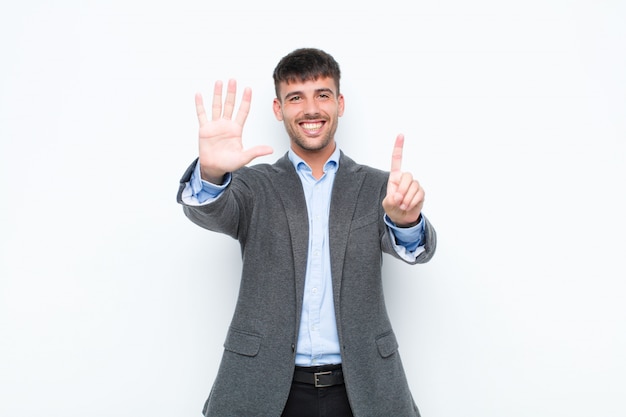 Jeune bel homme souriant et sympathique, montrant le numéro six ou sixième avec la main vers l'avant, compte à rebours contre le mur blanc