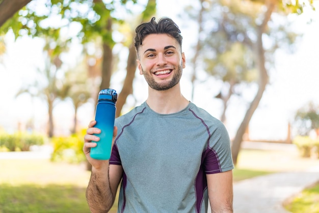 Jeune bel homme sportif avec une bouteille d'eau à l'extérieur souriant beaucoup