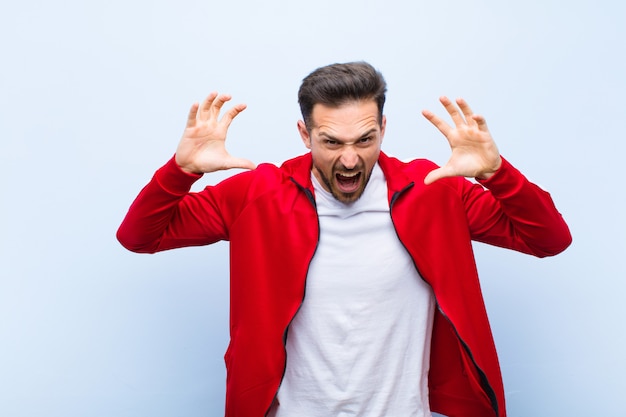 Jeune bel homme sportif ou moniteur criant de panique ou de colère, choqué, terrifié ou furieux, les mains à la tête contre le mur plat