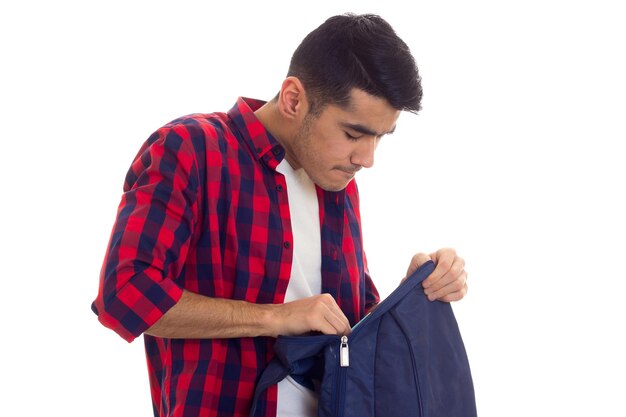 Jeune bel homme en T-shirt blanc et chemise à carreaux rouge regardant dans son sac à dos bleu