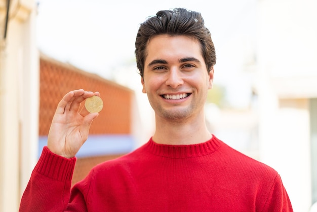 Jeune bel homme tenant un Bitcoin à l'extérieur souriant beaucoup