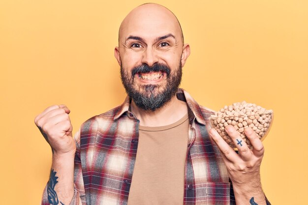 Jeune bel homme tenant un bol de pois chiches criant fier, célébrant la victoire et le succès très excité avec le bras levé