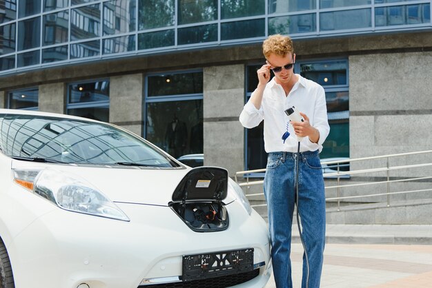 Jeune bel homme tenant un câble de charge au point de la station de charge électrique debout près de sa nouvelle voiture, l'air satisfait.