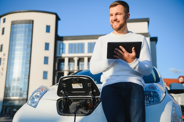 Jeune bel homme tenant un câble de charge au point de la station de charge électrique, debout près de sa nouvelle voiture.