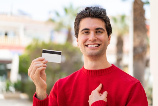 Jeune bel homme tenant une carte de crédit à l'extérieur avec une expression faciale surprise