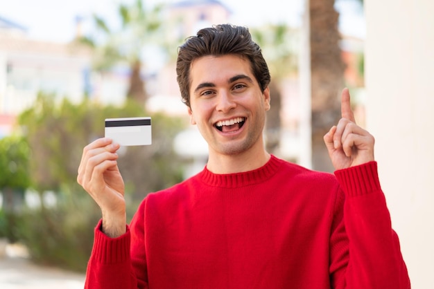 Jeune bel homme tenant une carte de crédit à l'extérieur pointant vers le haut une bonne idée