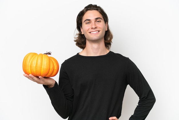 Jeune bel homme tenant une citrouille isolé sur fond blanc posant avec les bras à la hanche et souriant