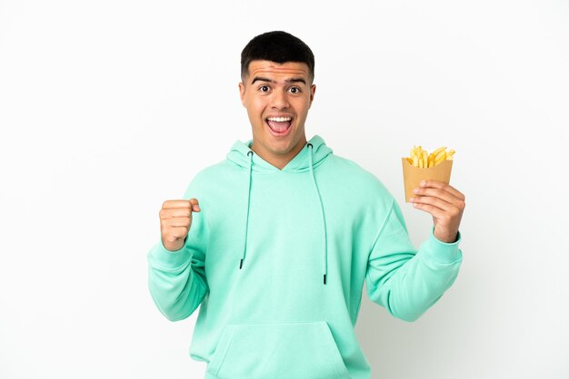 Jeune bel homme tenant des frites sur fond blanc isolé célébrant une victoire en position de vainqueur