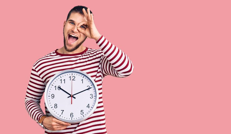 Photo jeune bel homme tenant une grande horloge souriant heureux faisant signe ok avec la main sur les yeux regardant à travers les doigts