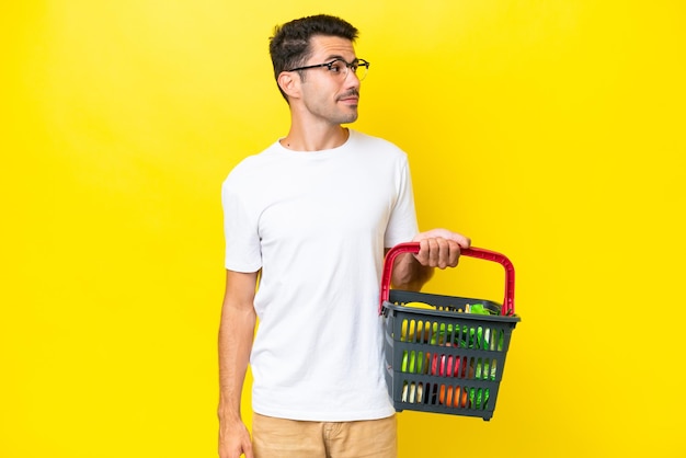 Jeune bel homme tenant un panier plein de nourriture sur fond jaune isolé regardant sur le côté