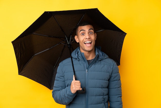 Jeune bel homme tenant un parapluie sur un mur jaune isolé avec surprise et expression faciale choquée