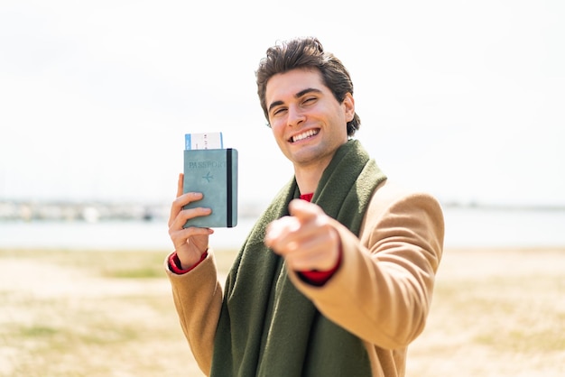 Jeune bel homme tenant un passeport à l'extérieur vous pointe du doigt avec une expression confiante