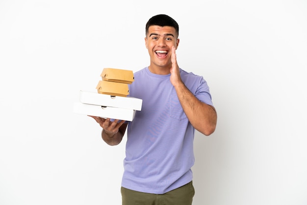 Jeune bel homme tenant des pizzas et des hamburgers sur fond blanc isolé avec une expression faciale surprise et choquée