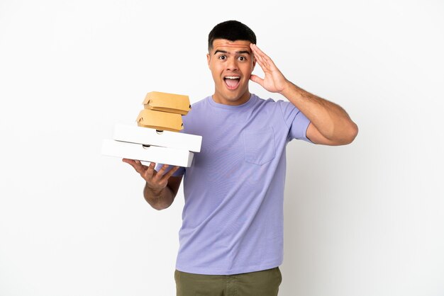 Jeune bel homme tenant des pizzas et des hamburgers sur fond blanc isolé avec une expression de surprise