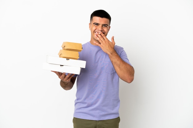 Jeune bel homme tenant des pizzas et des hamburgers sur fond blanc isolé heureux et souriant couvrant la bouche avec la main