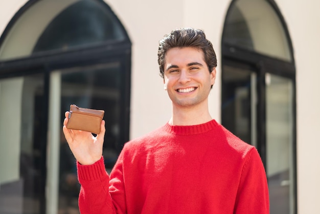 Jeune bel homme tenant un portefeuille à l'extérieur souriant beaucoup