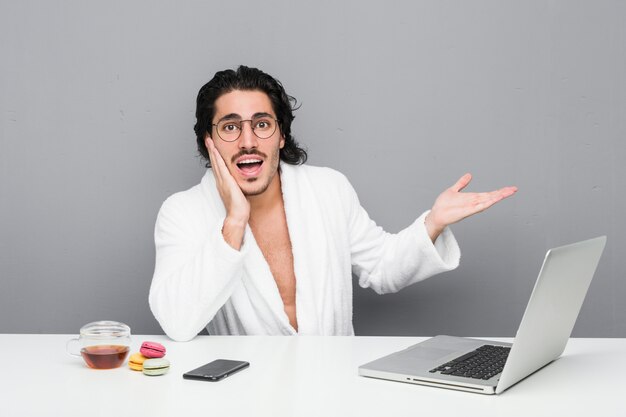 Jeune bel homme travaillant après une douche détient un espace de copie sur une paume, gardez la main sur la joue. Etonné et ravi.