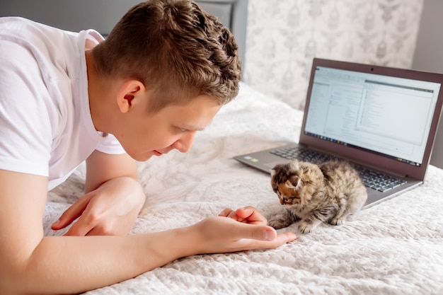 Jeune bel homme travaillant à la maison tout en mettant en quarantaine un coronovirus, couché avec un ordinateur portable et un chaton au lit