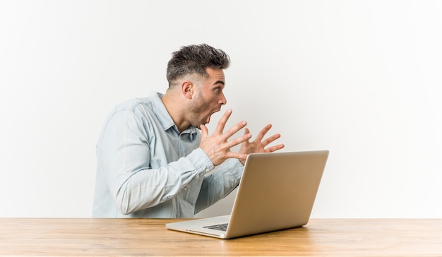 Jeune bel homme travaillant avec son ordinateur portable crie fort, garde les yeux ouverts et les mains tendues