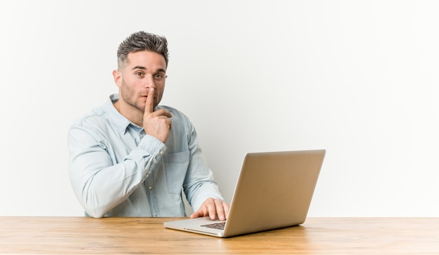 Jeune bel homme travaillant avec son ordinateur portable gardant un secret ou demandant le silence.