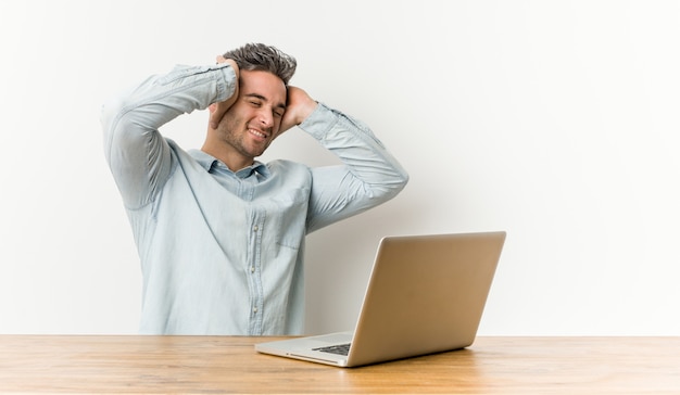 Jeune bel homme travaillant avec son ordinateur portable rit joyeusement en gardant les mains sur la tête.