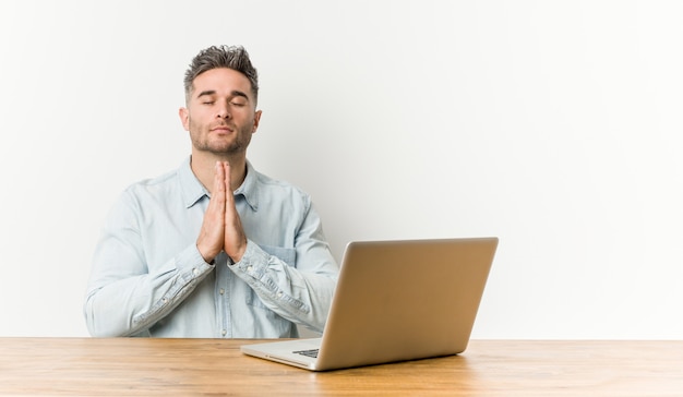 Jeune bel homme travaillant avec son ordinateur portable se tenant la main dans prier près de la bouche, se sent confiant.