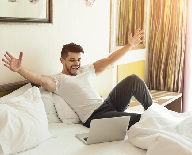 Photo jeune bel homme utilisant un ordinateur portable et célébrant le succès assis sur un lit dans une chambre d'hôtel