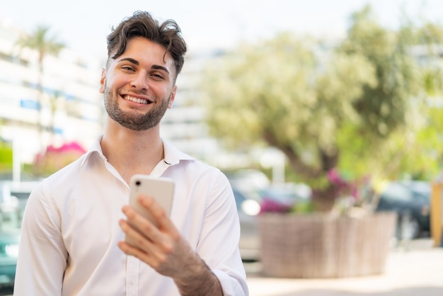 Jeune bel homme utilisant un téléphone portable à l'extérieur avec une expression heureuse