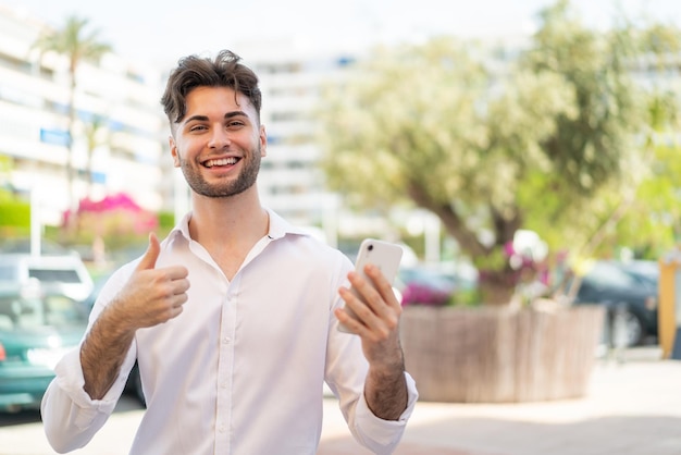 Jeune bel homme utilisant un téléphone portable à l'extérieur avec les pouces vers le haut parce que quelque chose de bien s'est produit