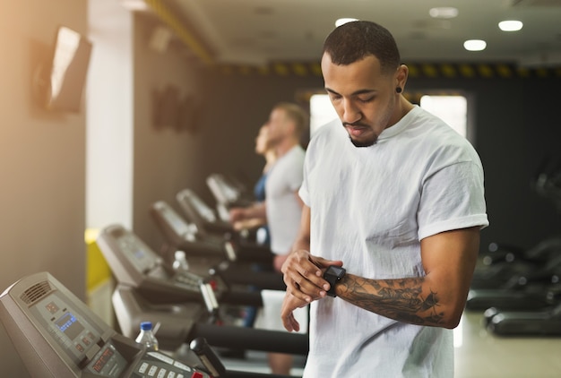 Jeune bel homme utilisant un tracker de fitness dans un club de sport. Entraînement cardio, course sur tapis roulant. Mode de vie sain, entraînement de gars en salle de sport, espace de copie