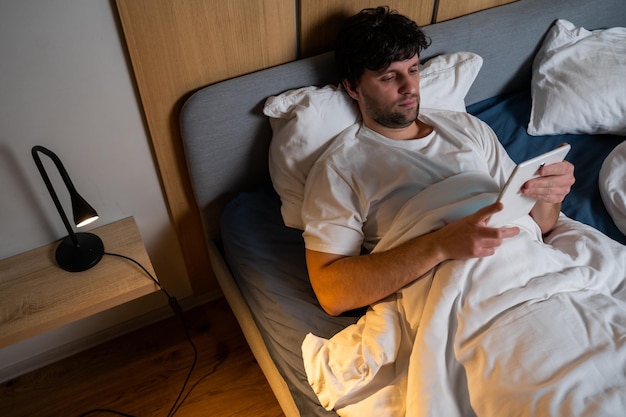 Jeune bel homme utilise une tablette allongée sur le lit la nuit Un homme souffrant d'insomnie utilise une tablette tout en se reposant sur un lit