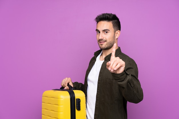 Jeune bel homme sur violet en vacances avec valise de voyage et en comptant un