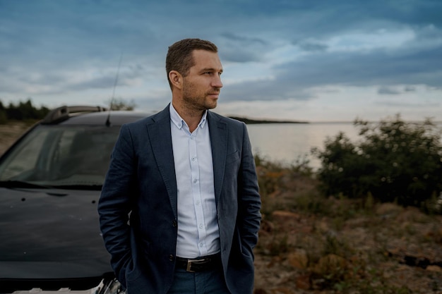 Jeune bel homme avec une voiture classique noire portant un costume noir. L'homme se tient près de sa voiture à l'extérieur.