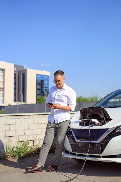 Jeune bel homme voyageant dans une voiture électrique s'arrêtant à une station de recharge debout branchant un câble naviguant sur Internet sur un smartphone souriant joyeusement tout en chargeant