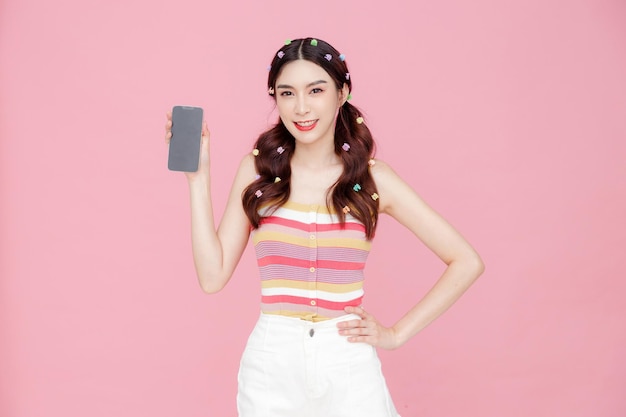 Jeune belle asiatique heureuse mignonne mince femme avec des nattes s'amusant et posant sur fond isolé rose Modèle féminin élégant montrant un téléphone à écran vide et les mains sur la taille en studio