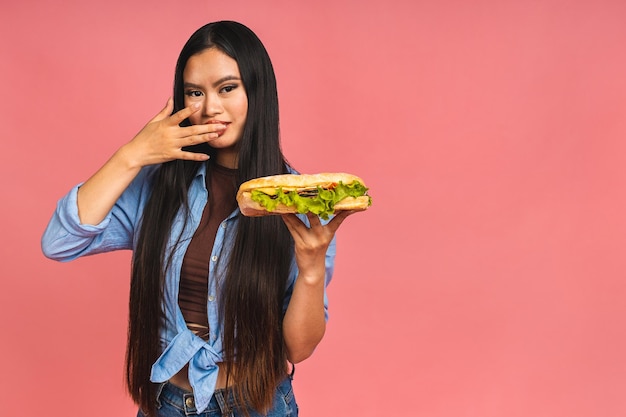 Jeune belle asiatique japonaise chinoise femme mangeant un sandwich ou un gros hamburger avec satisfaction Fille apprécie le concept de régime à emporter hamburger savoureux debout isolé sur fond rose