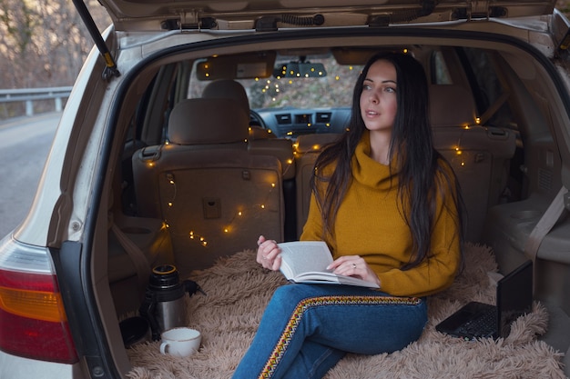 La jeune belle brune aime se détendre dans le coffre de sa voiture. Une guirlande jaune et un plaid beige créent le confort, Femme lisant un livre et buvant du thé chaud