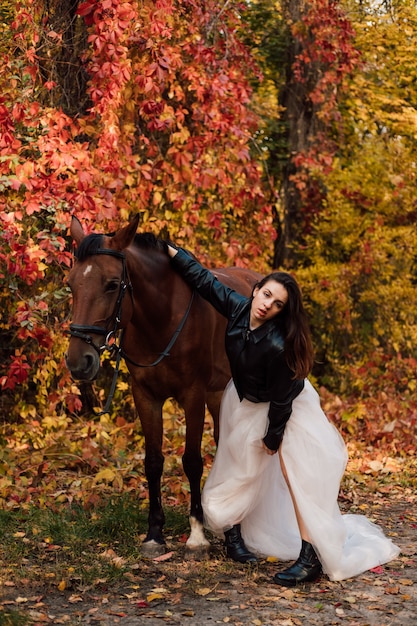 Jeune belle brune dans une robe blanche et une veste noire en cuir posant près d'un cheval