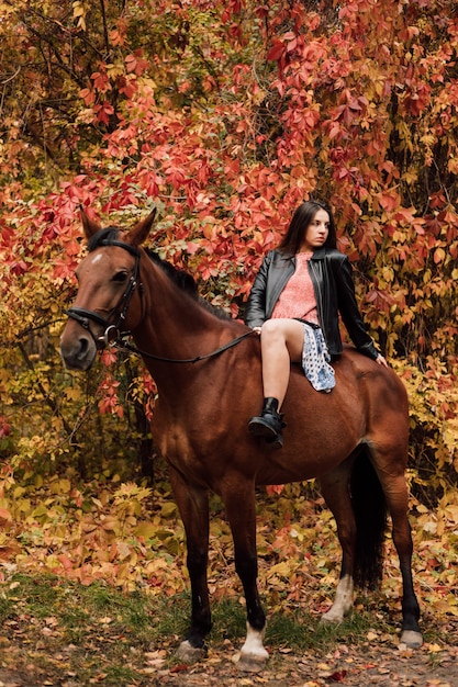 Jeune belle brune dans une robe et une veste noire en cuir sexy est assise sur un cheval